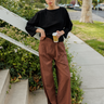 A woman stands on outdoor concrete steps beside a sidewalk, wearing a loose black quarter-sleeve top and brown wide-leg pleated trousers, holding a takeaway coffee cup.