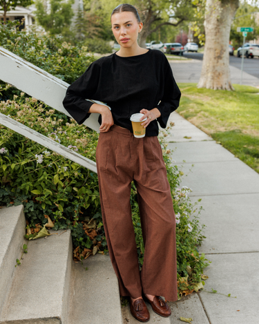 A woman stands on outdoor concrete steps beside a sidewalk, wearing a loose black quarter-sleeve top and brown wide-leg pleated trousers, holding a takeaway coffee cup.