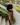 A woman stands on outdoor concrete steps beside a sidewalk, wearing a black quarter-sleeve top and brown wide-leg pants, holding a paper coffee cup. The background shows a residential street with trees, grass, and parked cars.