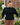 A woman wearing a black quarter-sleeve peplum-style blouse and brown plaid trousers, standing outdoors in front of a brick wall and greenery.