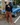 A woman stands on a street in front of a blue classic car, wearing a black quarter-sleeve top, brown wide-leg plaid pants, and brown loafers, while holding a dark blue denim jacket.