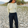 A woman standing barefoot on a rocky outdoor riverbed wearing a pale green pleated blouse, dark blue jeans with rolled cuffs, and a cream knit beanie.