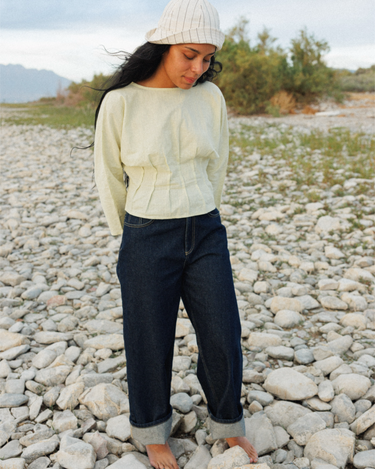 A woman standing barefoot on a rocky outdoor riverbed wearing a pale green pleated blouse, dark blue jeans with rolled cuffs, and a cream knit beanie.