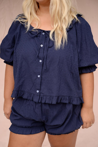 Woman wearing a navy eyelet, button-up blouse with a ruffled neckline and matching bloomer-style shorts in front of a pink background.