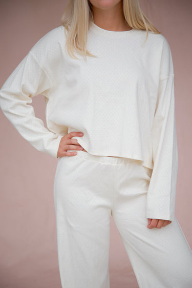 Woman wearing a cream, long-sleeve pointelle top and matching pointelle pants against a pink background.