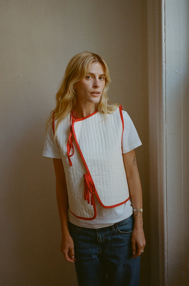 Woman standing in front of a white wall wearing a cream vest with red trim over a white short sleeve top, paired with medium wash jeans.