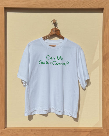 White t-shirt with 'Can My Sister Come?' in green text on a wooden hanger inside a wooden frame.
