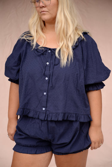 Woman wearing a navy eyelet, button-up blouse with a ruffled neckline and matching bloomer-style shorts in front of a pink background.