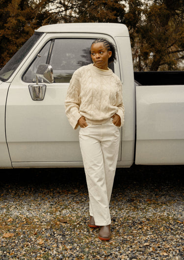 Woman wearing a cream sweater and white denim jeans in front of a vintage white truck outdoors.