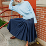 A woman wearing a light blue knit sweater and a navy pleated midi skirt sits on concrete steps beside a brick building, with a red railing and a coffee cup visible in the background.