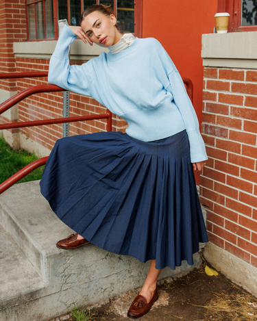 A woman wearing a light blue knit sweater and a navy pleated midi skirt sits on concrete steps beside a brick building, with a red railing and a coffee cup visible in the background.