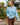 A woman wearing a light blue crewneck knit sweater and brown plaid pants walks outdoors while holding a paper coffee cup.