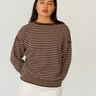 A woman modeling a brown and white horizontally striped knit sweater with a crew neck, paired with white pants, against a plain light gray background.