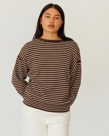 A woman modeling a brown and white horizontally striped knit sweater with a crew neck, paired with white pants, against a plain light gray background.