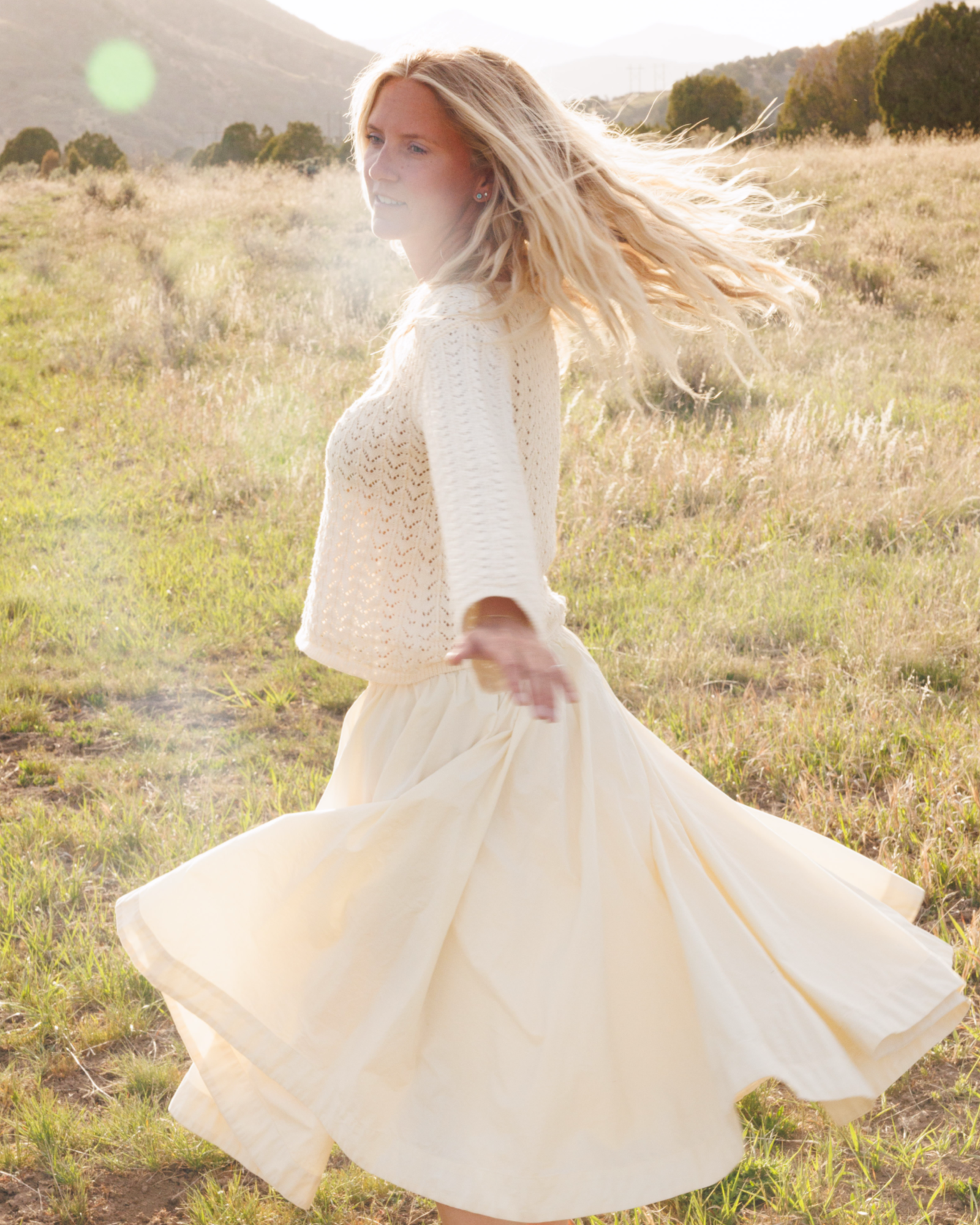 Woman in a crochet knit sweater and cream-colored skirt spinning in a field with mountains in the background.