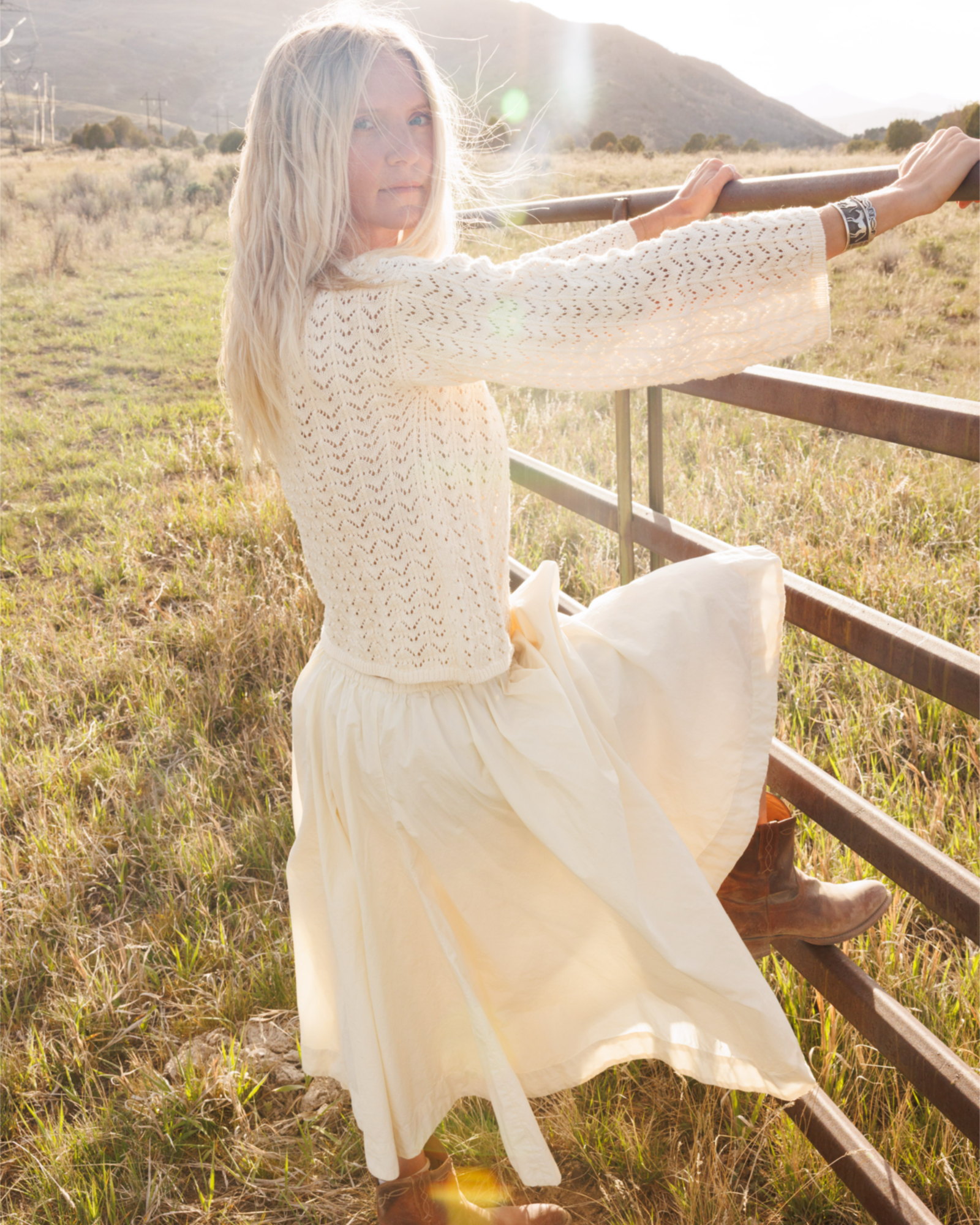 Woman in a cream outfit standing by a wooden fence in a field with mountains in the background