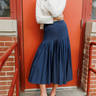 A person wearing a navy blue pleated midi skirt, a white long-sleeve mock-neck shirt, and brown leather loafers, standing in front of a red-orange brick door.