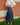 A person wearing a cream long-sleeve top and a dark navy pleated maxi skirt, standing outdoors on grass in front of a red brick wall.