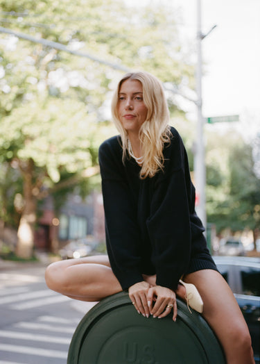 Woman sitting on a green mailbox outside in an urban setting wearing a black knit sweater with matching knit shorts.